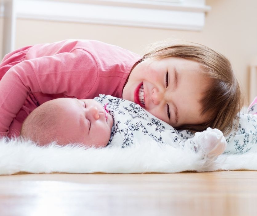 A smiling young toddler in a pink shirt leans over a sleeping newborn baby lying on a white faux-fur rug.