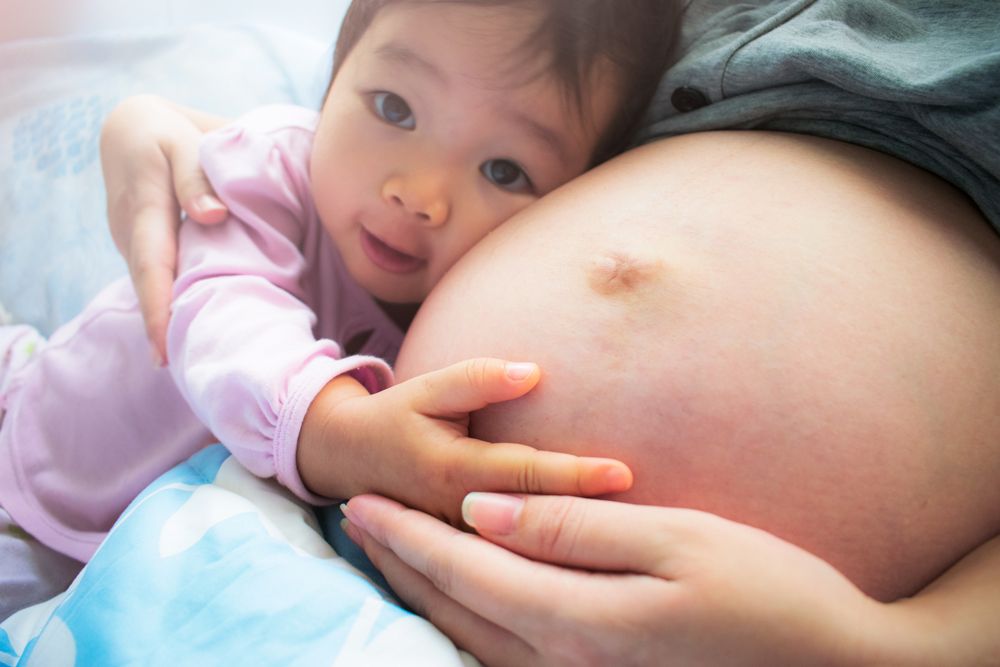 Close-up of a young child leaning their head against a pregnant person's bare stomach while touching their belly.