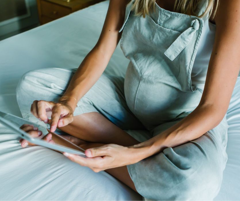 A pregnant person in light green overalls sits cross-legged on a bed while using a tablet.