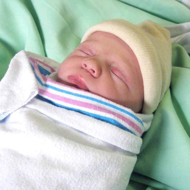 A sleeping newborn baby wearing a yellow knit cap, swaddled in a pink and blue striped hospital blanket.