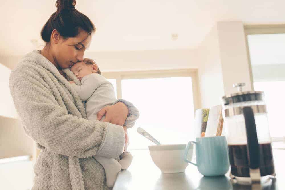 Mother in a gray robe holding a sleeping baby in a kitchen near a French press and coffee mug.
