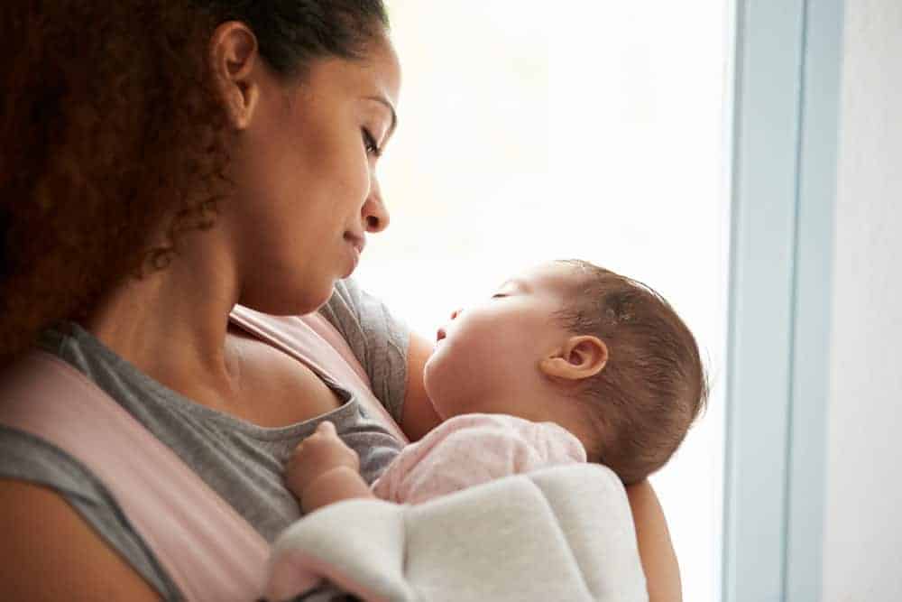 A woman looks down affectionately at a sleeping infant held in her arms against a bright background.