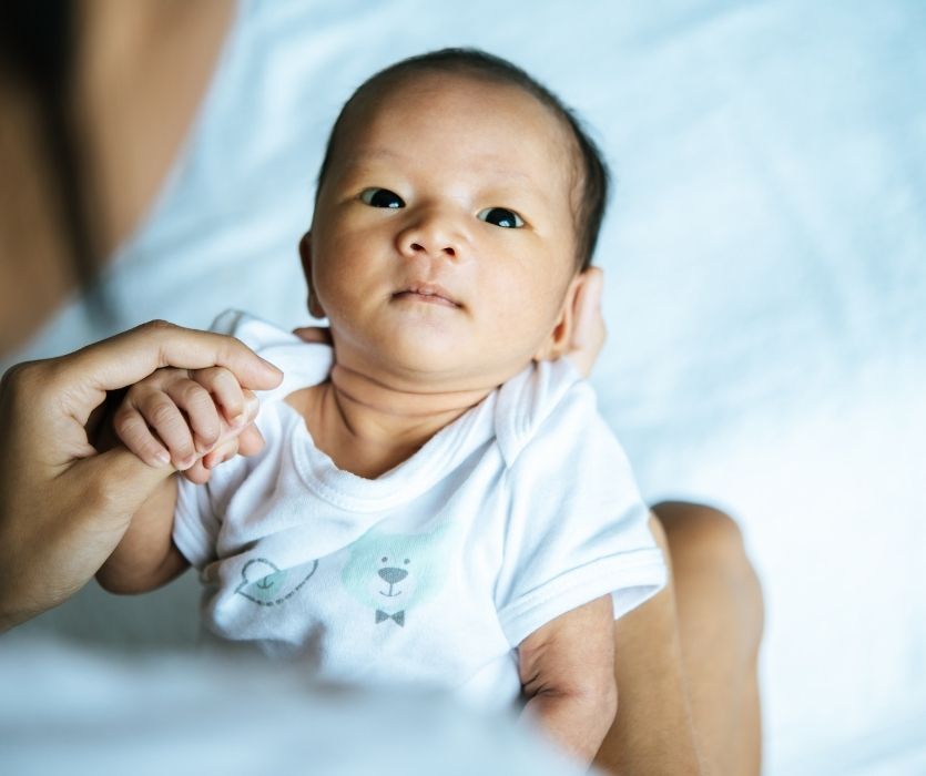 A close-up of a wide-eyed newborn baby in a white shirt, being held and having their hand grasped by an adult.