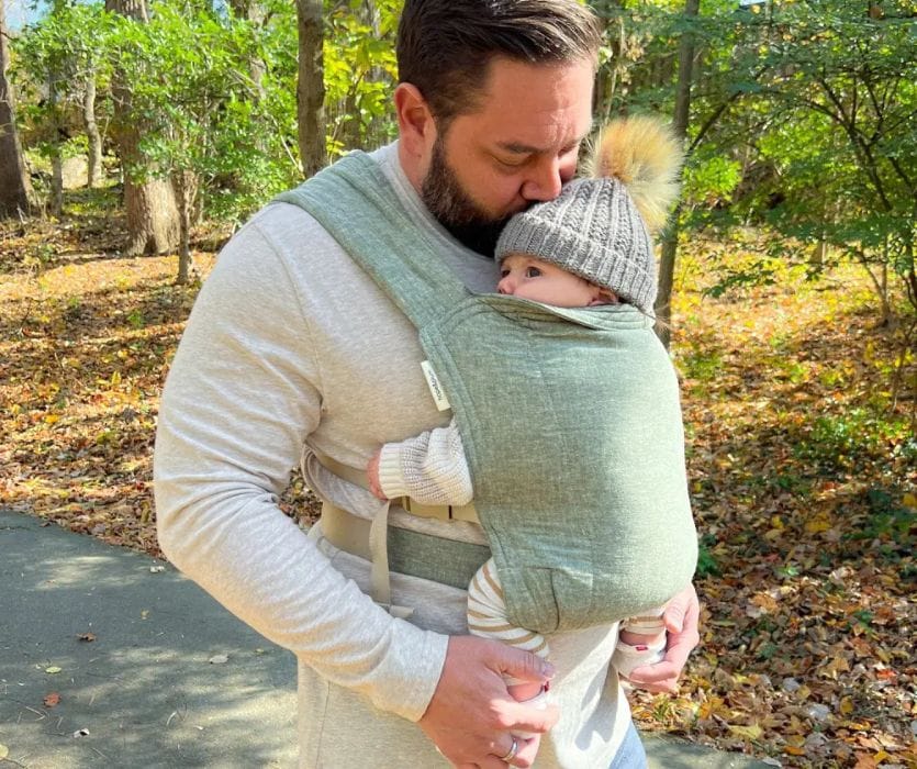 A man kissing a baby’s head while carrying them in a green fabric wrap baby carrier outdoors.