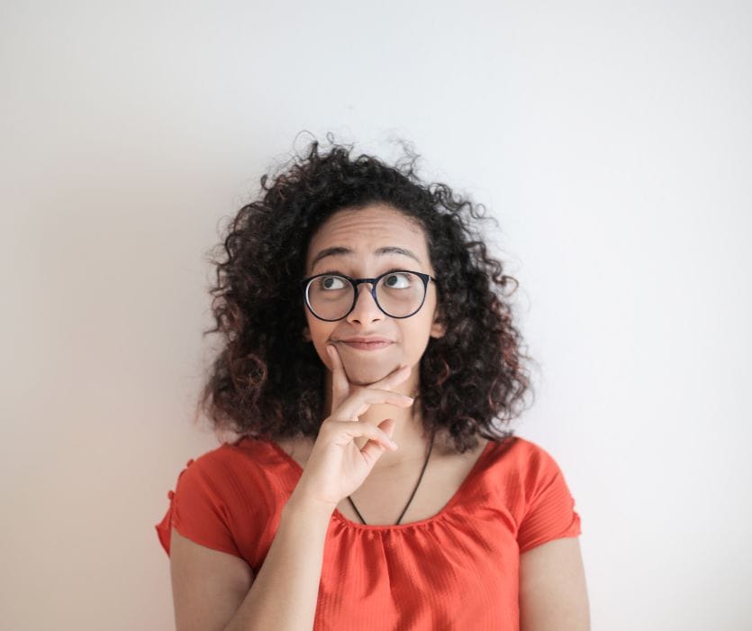 A person with curly hair and glasses looking upward with a thoughtful expression and a hand on their chin.