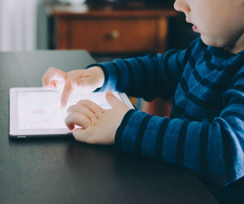 A young child in a blue striped shirt uses their fingers to touch a glowing tablet on a dark table.