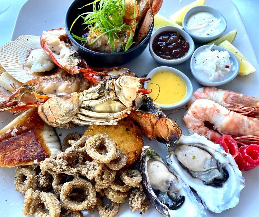 A platter of lobster, shrimp, oysters and calamari sits with lemon wedges and sauces beside guidance on safe seafood and fish during pregnancy.