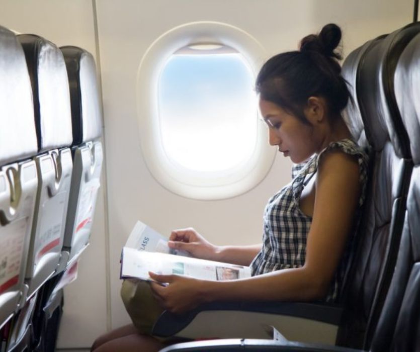 Woman sitting in an airplane seat by a window reading a magazine.