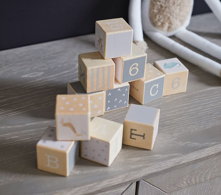 Neutral-toned wooden baby blocks featuring letters, numbers, and illustrations stacked on a grey wood surface.