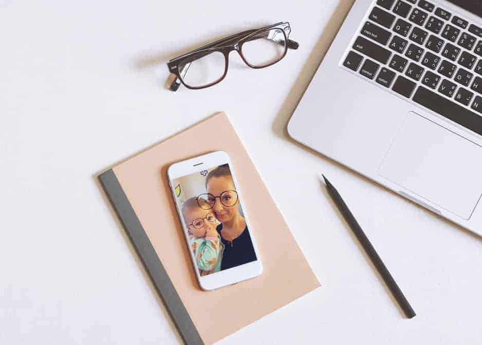 Smartphone showing a photo of a mother and baby on a desk with a laptop, notebook, glasses, and pencil.
