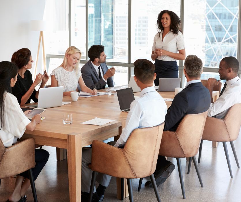 A professional woman leads a meeting with a diverse group of colleagues seated around a large wooden boardroom table.