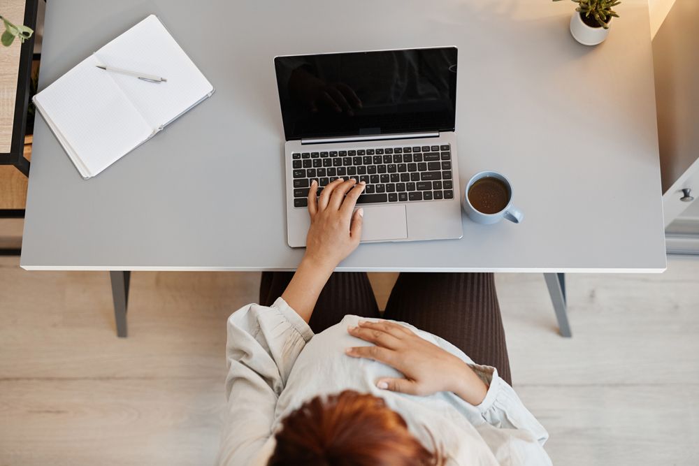 Overhead view of a pregnant woman sitting at a desk, typing on a laptop and resting her hand on her stomach.