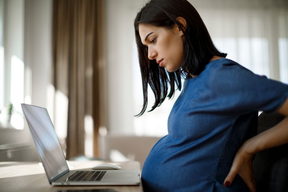 A pregnant woman sits at a desk looking at a laptop while holding her lower back in discomfort.